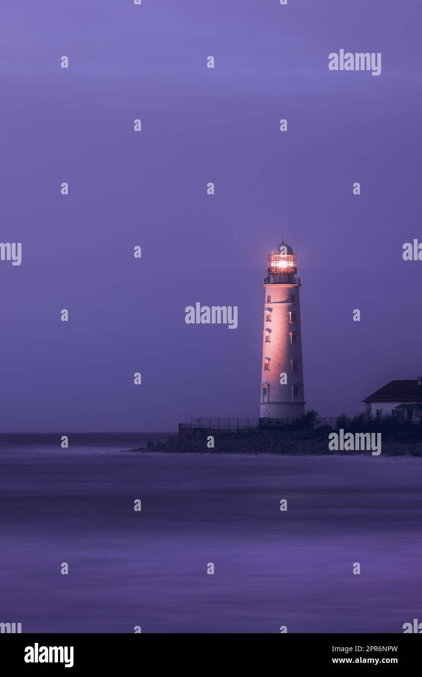 A lighthouse on the coast of the sea at dusk, its bright lamp is a landmark for ships. Seascape, nature background. Stock Photo