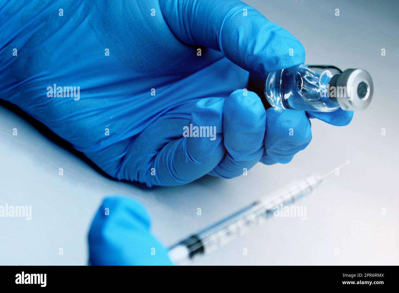 Close-up of a hand in a blue medical seal holding a vial of a vaccine. In the foreground, the syringe is out of focus. The concept of the fight against Covid19. Stock Photo