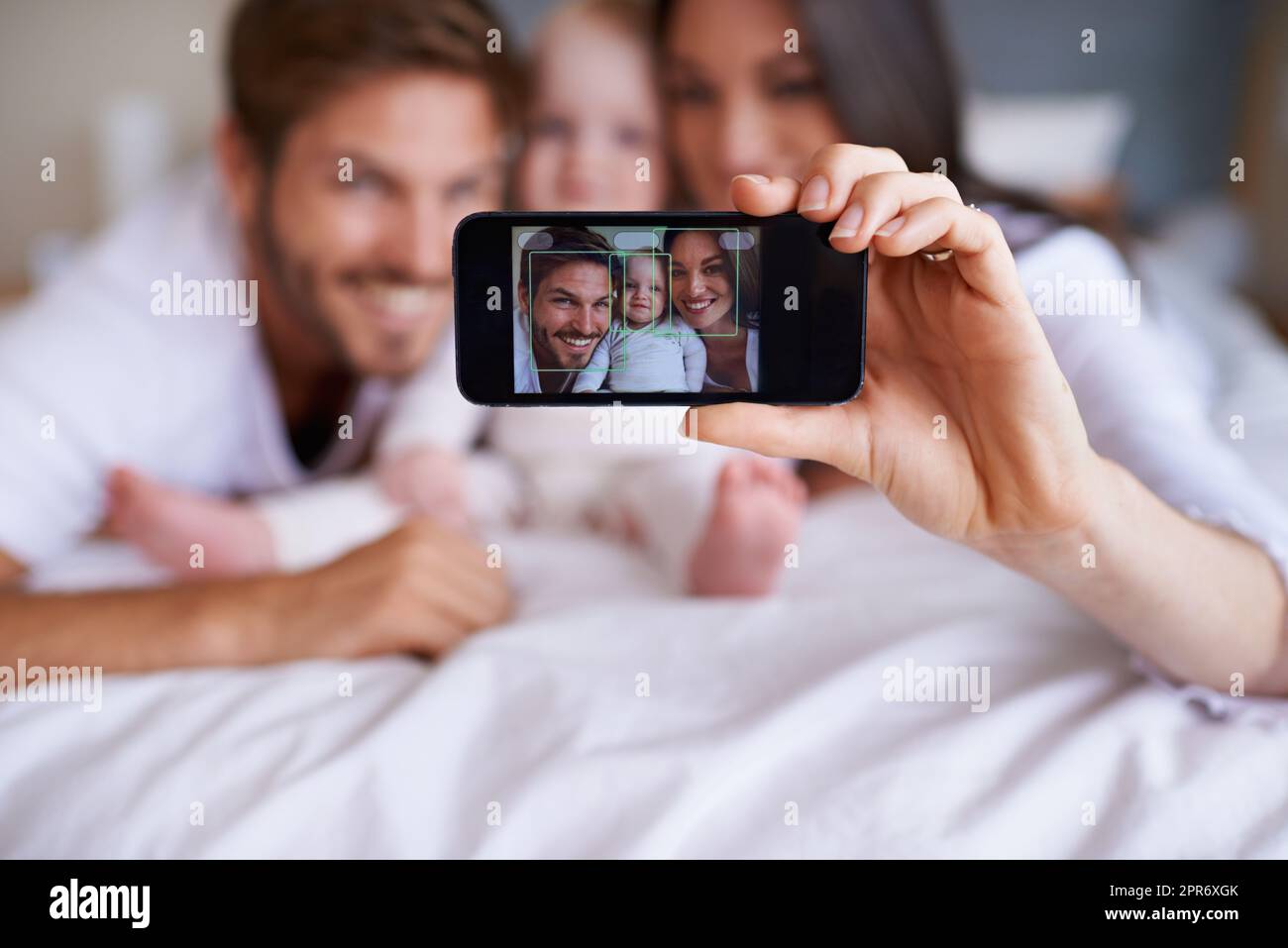 Smile for the family selfie. Shot of a young couple lying on the bed with their baby girl and taking a selfie with their mobile phone. Stock Photo