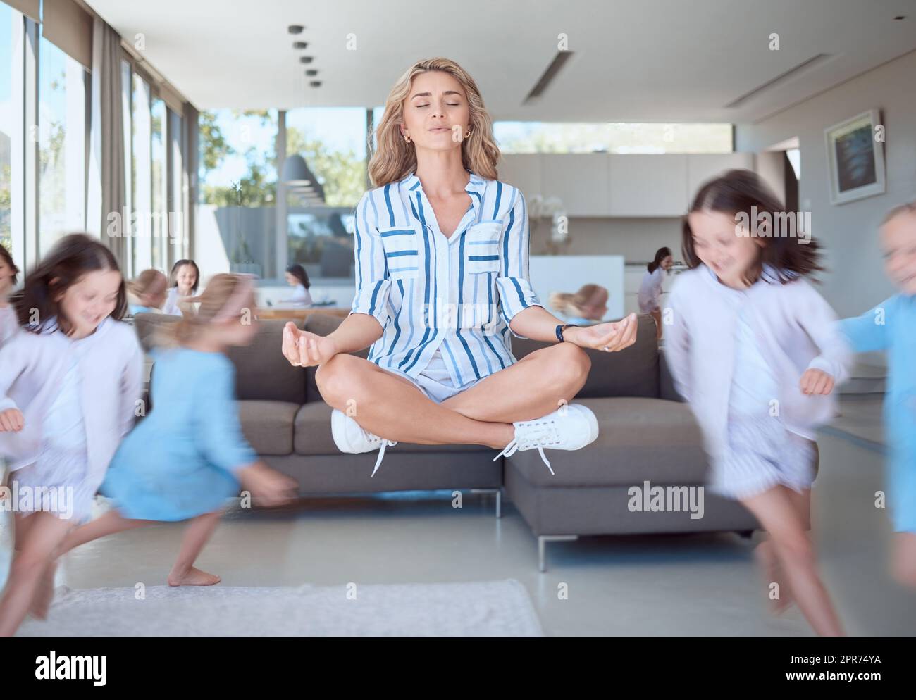 Young mother meditating for stress relief while her energetic kids run around her. Calm mom with naughty mischievous children running around her in the living room at home Stock Photo