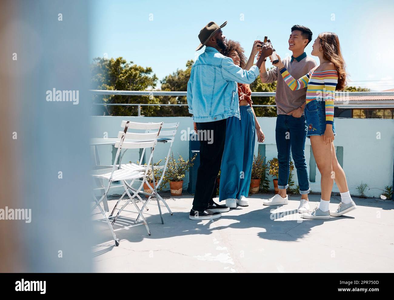 Full length of diverse group of young millennial friends standing outside together and toasting with beers. Smiling men and women being social and celebrating on a rooftop. Weekend party and alcohol Stock Photo