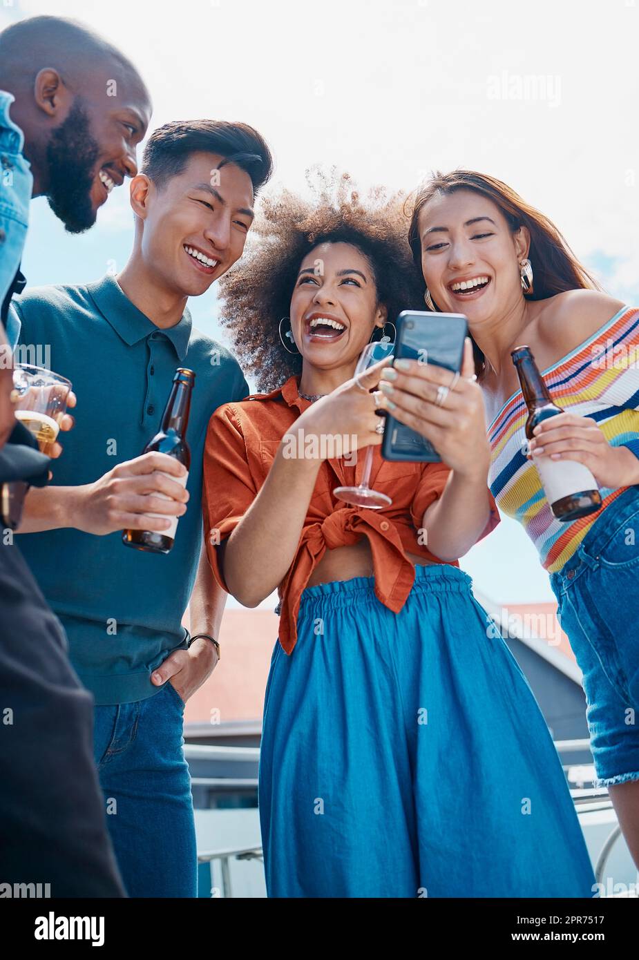 A group of diverse young cheerful friends standing outside together and using a cellphone for selfies and social media. Smiling men and women being social and celebrating on a weekend party Stock Photo