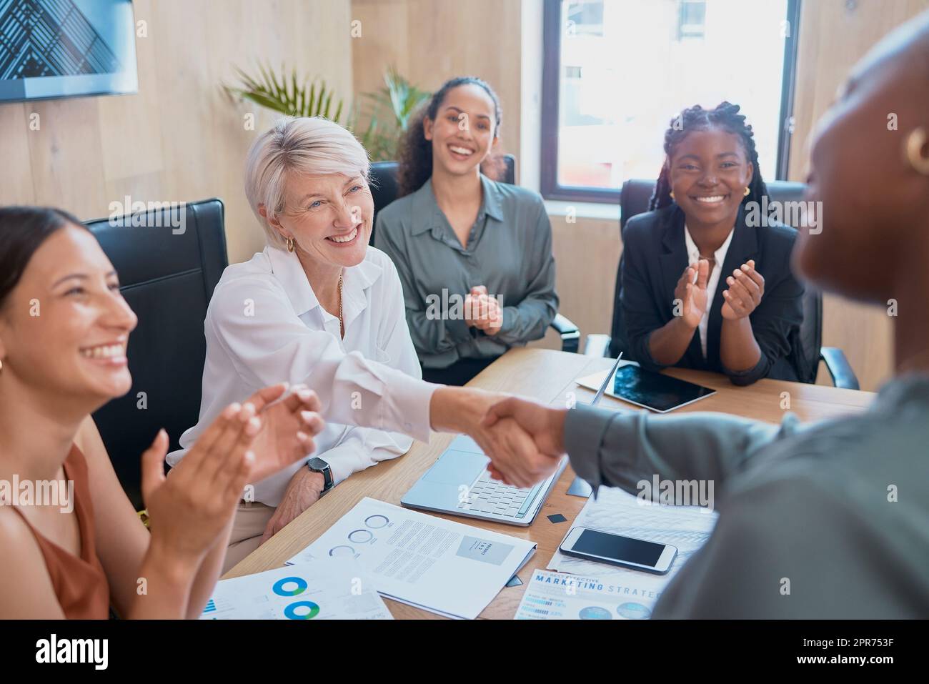 Handshake between business women. Two diverse colleagues shaking hands during a meeting in the boardroom. Congratulating her on a job well done while coworkers clap in acknowledgement of achievement Stock Photo