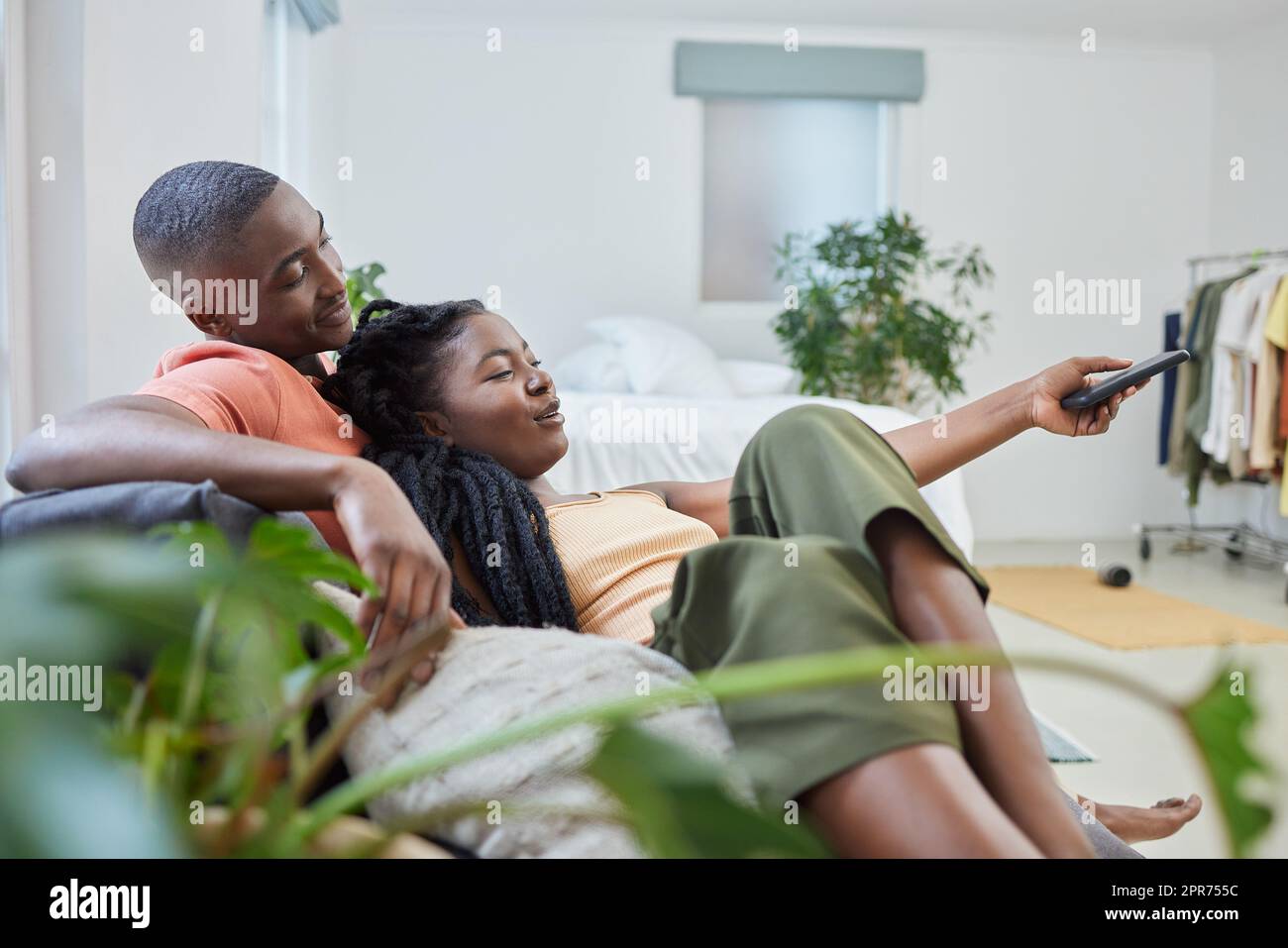 Young african american couple changing channels on remote and watching television together on sofa at home. Girlfriend relaxing on boyfriends lap while enjoying entertainment shows, series and movies Stock Photo