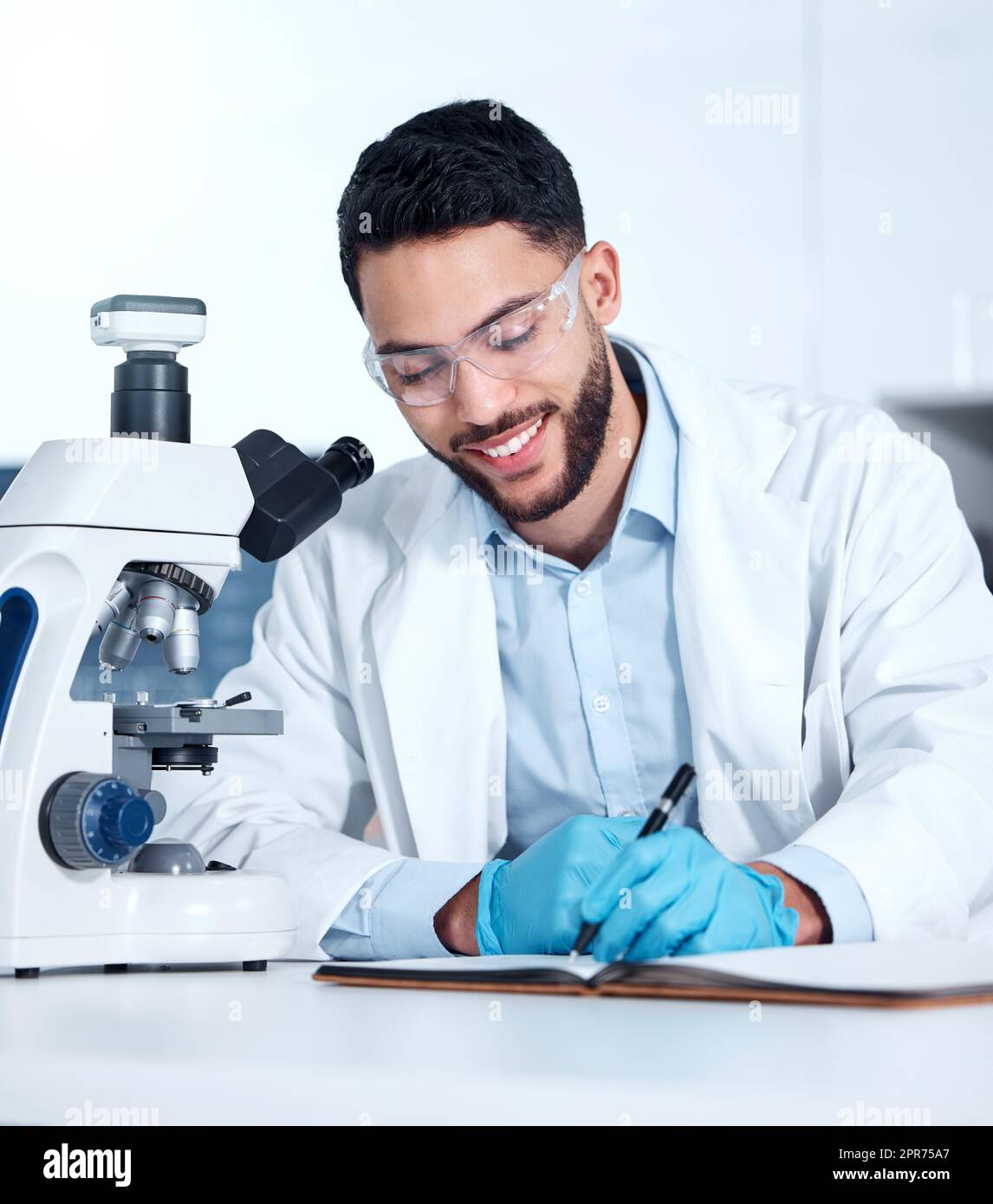 One handsome young mixed race man wearing gloves and a labcoat and looking at medical samples on a microscope in a lab. A male scientist wearing safety goggles and smiling while writing in a notebook Stock Photo