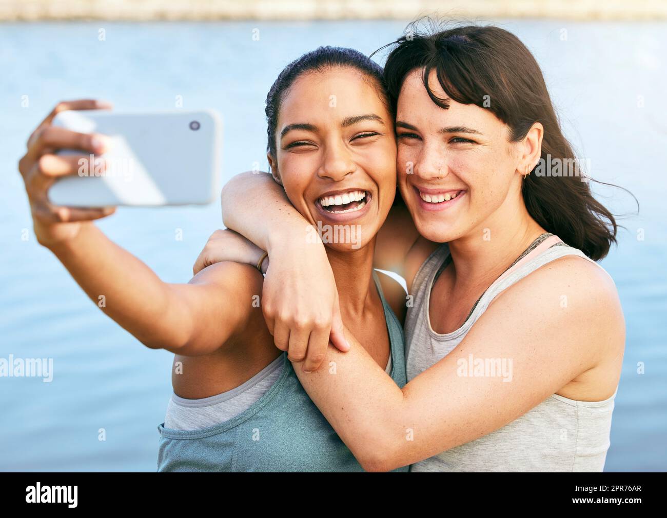 Two female friends taking selfies after a workout in nature and standing in front of a lake using a smartphone. Young Latino female using her phone to take a selfie of herself and her friend Stock Photo