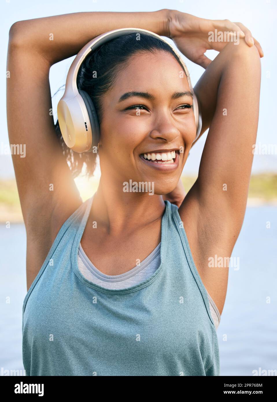 One active latina woman stretching arms and triceps by pulling elbow towards spine while exercising outdoors. Female athlete doing warm up to prepare body and muscles for training workout or run while listening to music on wireless headphones Stock Photo
