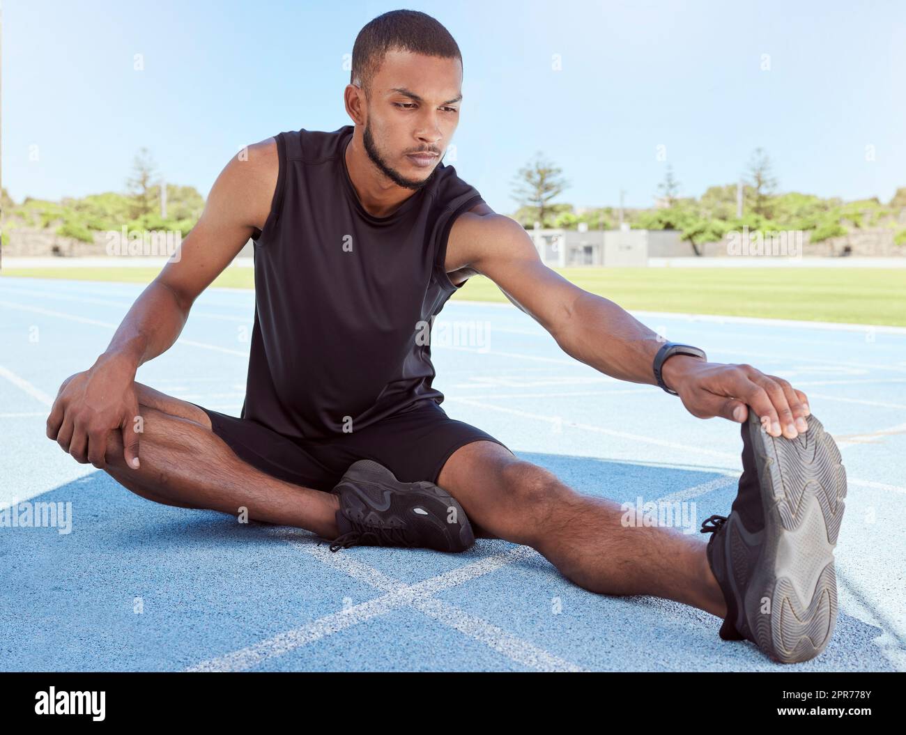 Male runner doing stretching exercises while sitting on sports track. Determined young male athlete sitting alone and warming up his muscles before going for a run. Fitness man preparing for a race Stock Photo