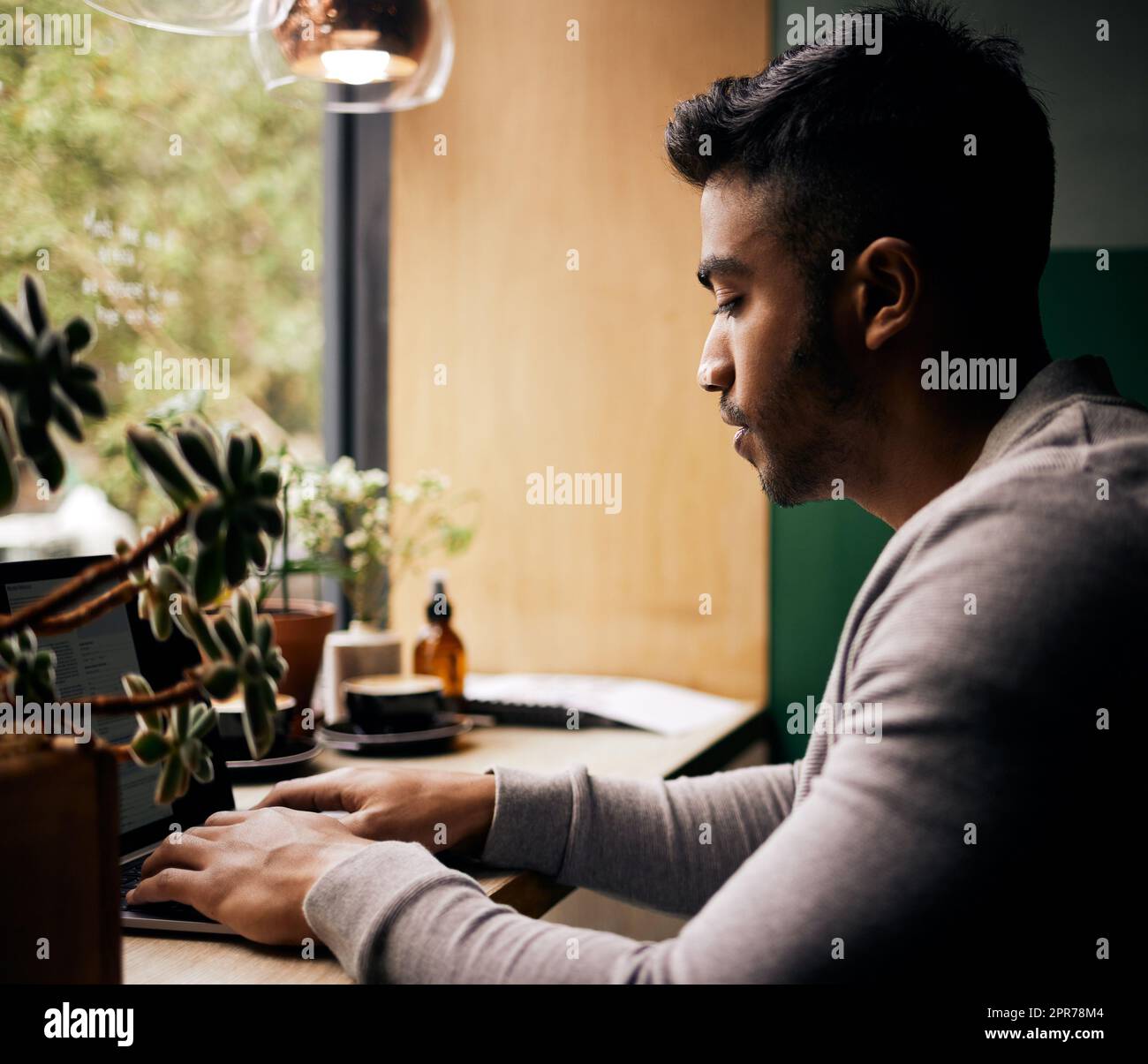 Young mixed race man or student browsing on a laptop in a cafe. One indian guy studying or working freelance at a restaurant. Typing to do research and planning online for blog, work or assignment Stock Photo