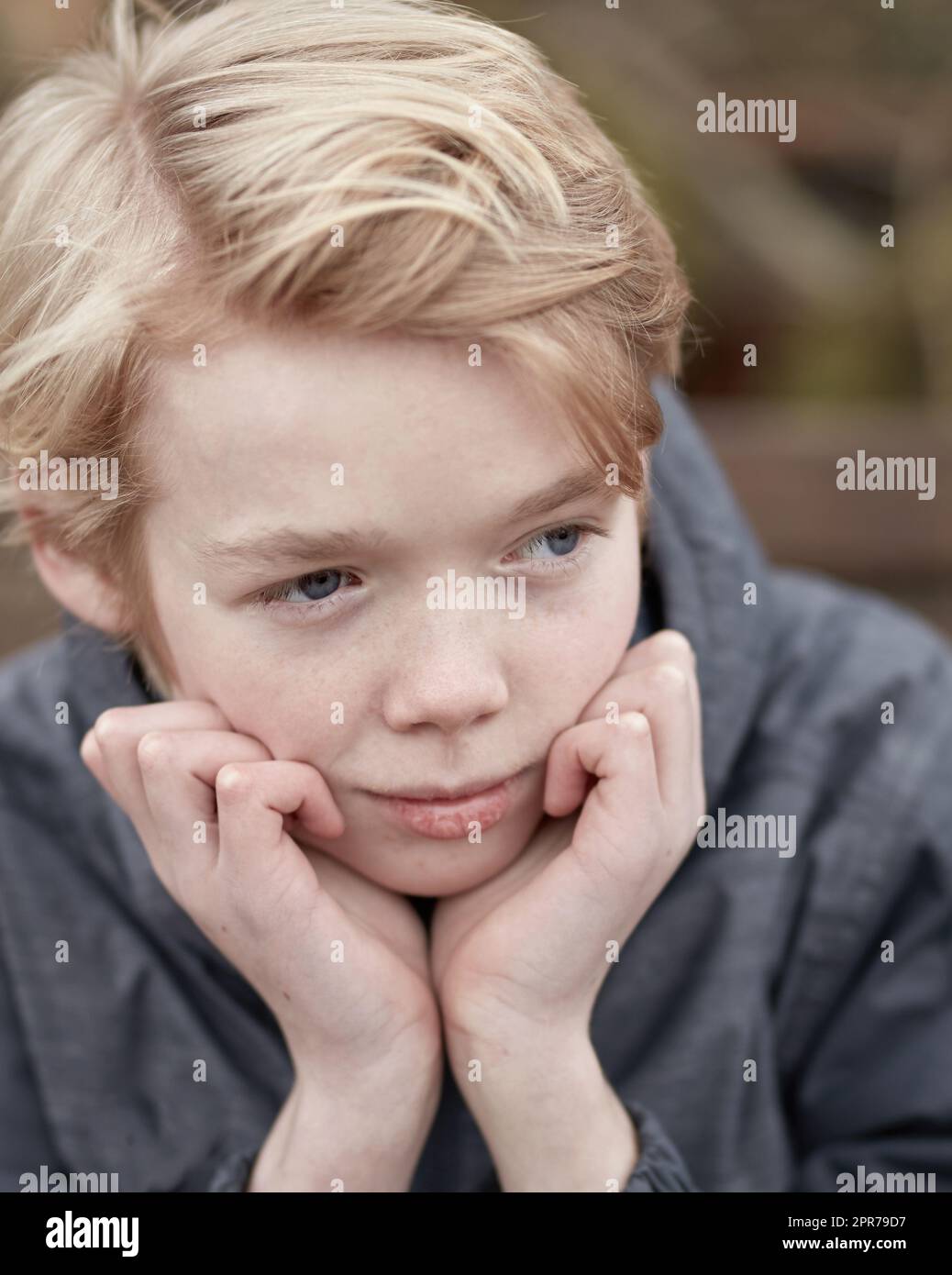 Lonely kid with his hands on his chin thinking. Young little teen kid outside on a playground alone. Cute caucasian male child serious. Bored teenage boy face sitting outdoors in a park alone. Stock Photo