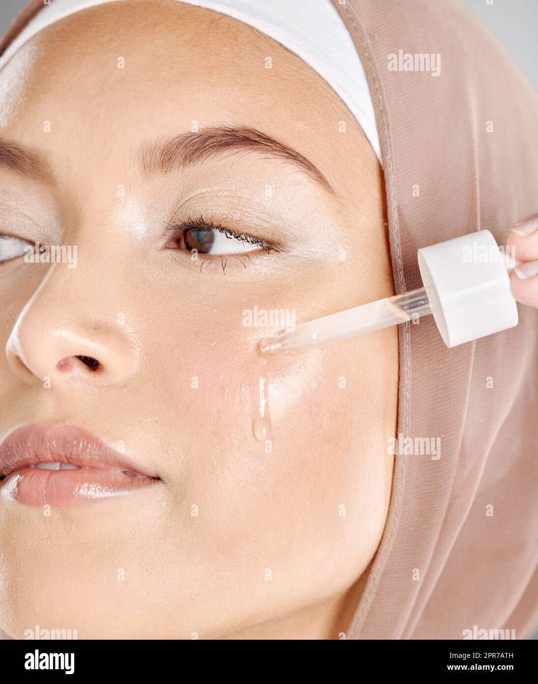 A young woman applying antiaging facial serum to her face and skin. Beautiful muslim girl wearing a hijab trying retinol with a dropper as her skincar Stock Photo