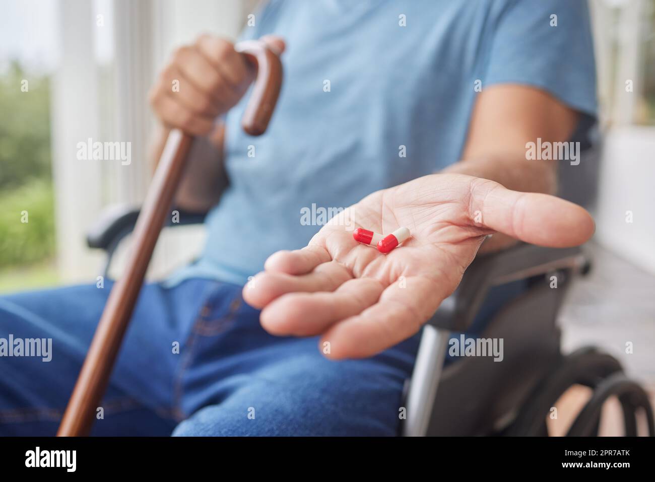 Sick senior man in recovery holding his medication, sitting in a wheelchair using a walking stick in a nursing home. Hand of a mature man taking his daily treatment for chronic disease and illness Stock Photo