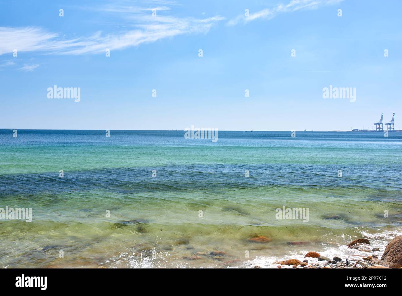 Landscape view of a blue ocean with clear skies on the horizon on a warm summer day. Little waves crash on a quiet calm rocky shore in the mediterranean sea. Panoramic coastline location for tourist Stock Photo