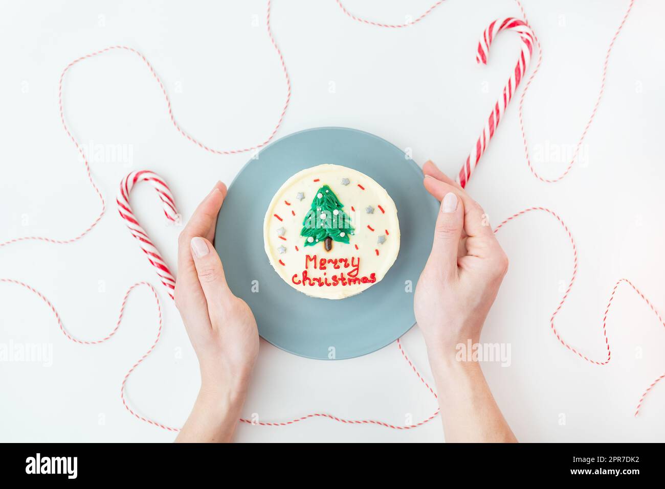 A girl holds a plate with a Christmas cake with the inscription Merry Christmas, decorated with icing, on a white background with lollipops and a red thread for gifts. Stock Photo
