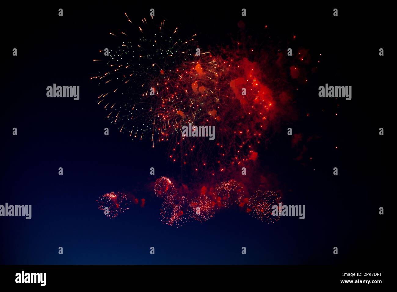 Red and orange fireworks with green sparks inside and smoke, below you can see small fireworks against the night sky Stock Photo