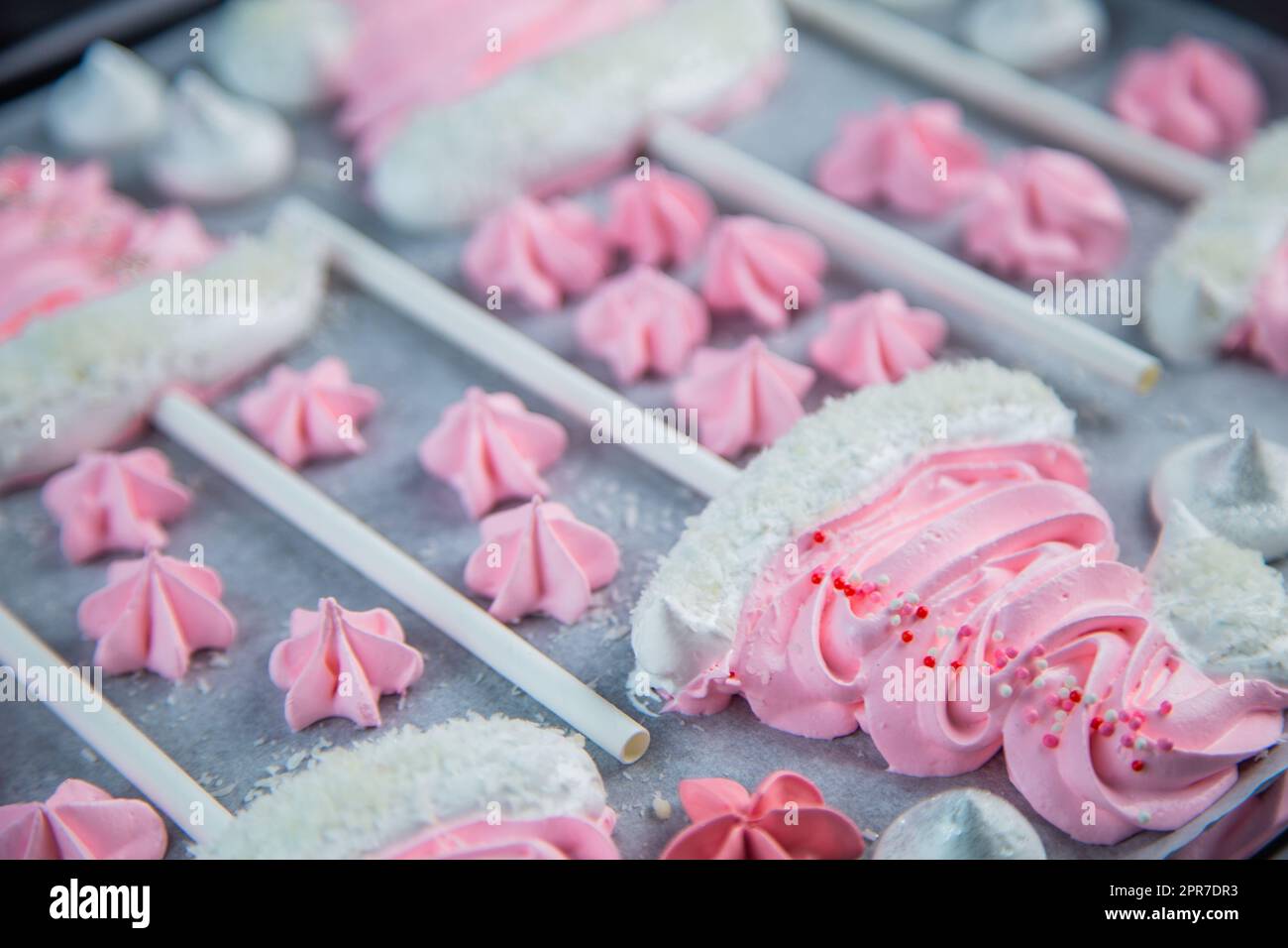 Meringues in the form of pink Christmas hats with a pompom and a white lapel on a stick lie on white parchment in two rows, pink stars and white meringues lie between them Stock Photo