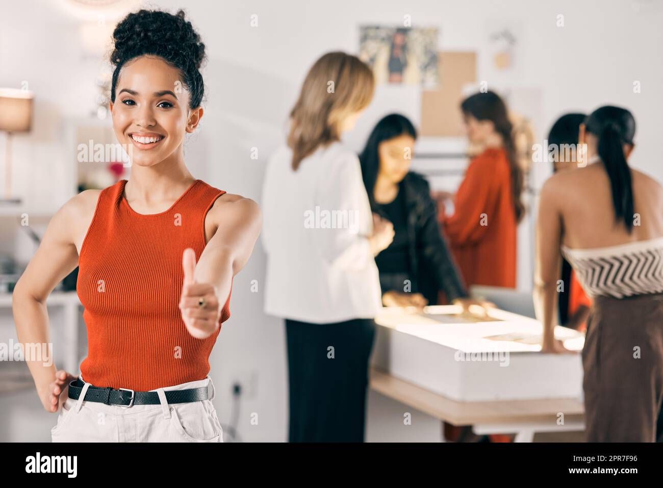 I aim to please. Shot of a young woman showing the thumbs up while her coworkers collaborate in the background. Stock Photo