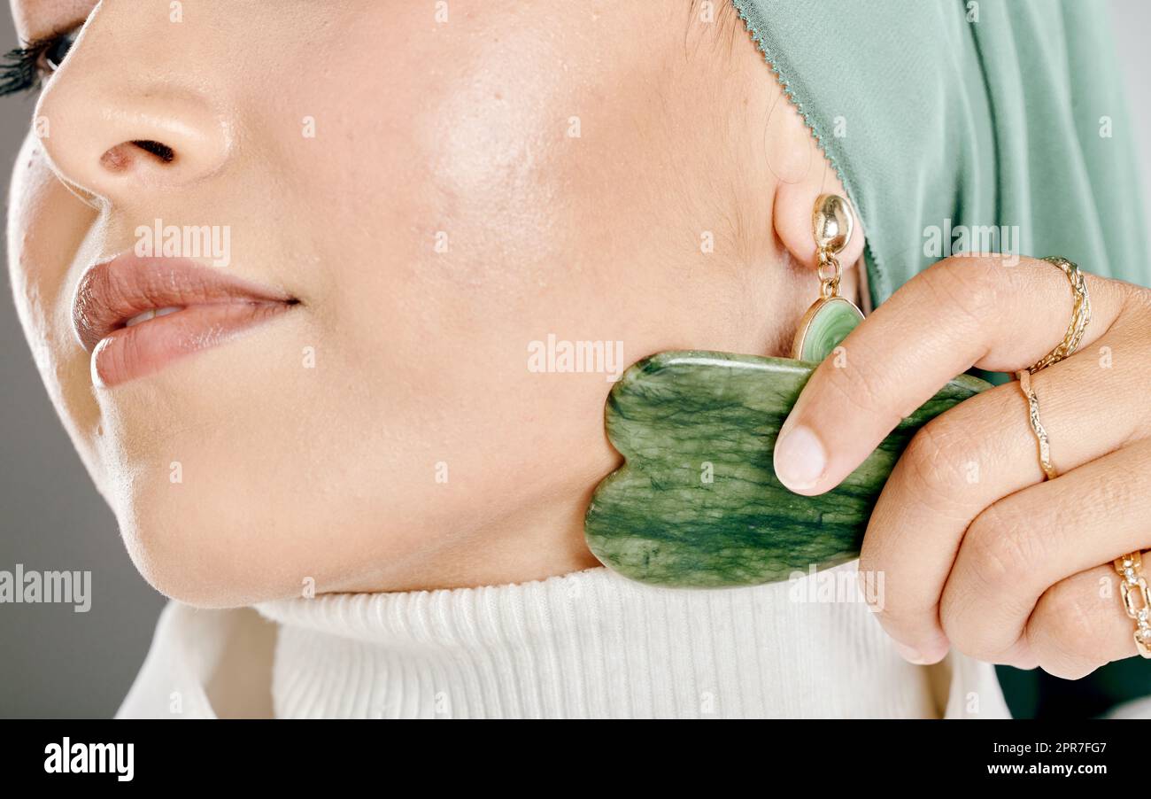 Gua sha massage on young womans face. Closeup of a muslim woman wearing a hijab or headscarf while using an anti ageing tool to reduce the appearance of wrinkles, puffiness and promote cell renewal Stock Photo