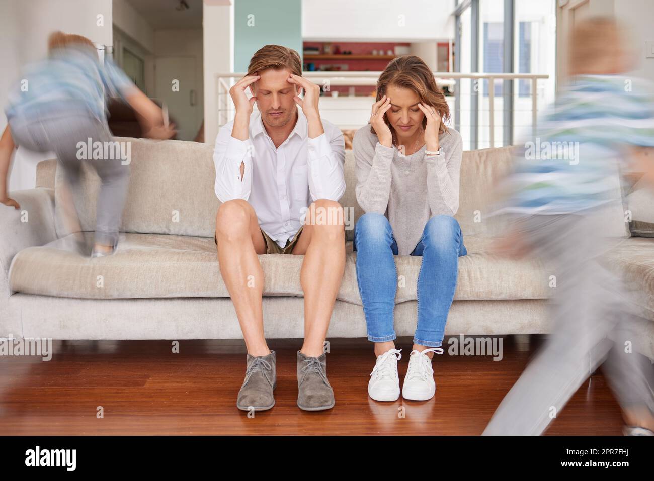 Naughty hyperactive, energetic children annoying their mother and father at home. Tired and stressed parents suffering from a headache and fatigue while adhd kids run around living room being noisy. Stock Photo