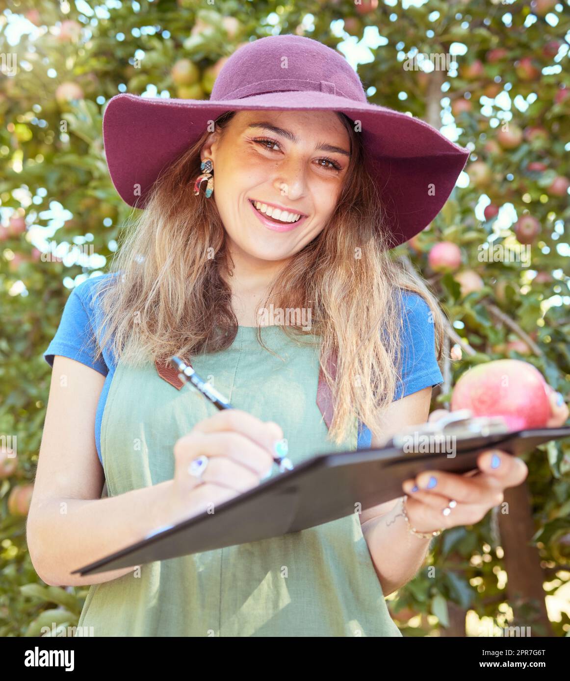 Portrait of female farm worker holding an apple while writing and making notes on a orchard farm during harvest season. Agronomist doing inspection and record keeping on the quality of produce Stock Photo