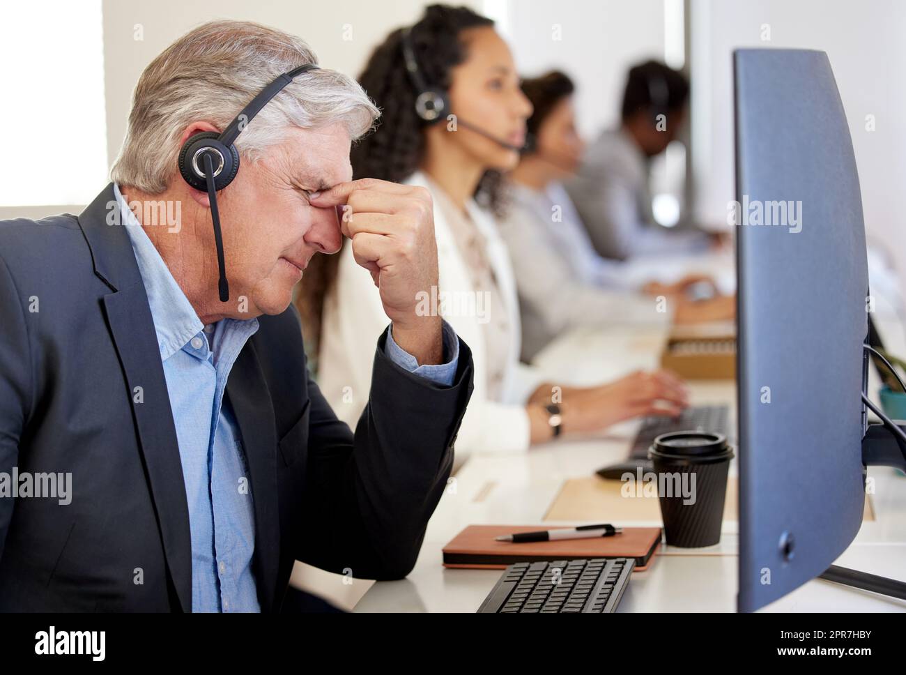 The solution is nowhere in sight. a mature call centre agent looking stressed out while working in an office with his colleagues in the background. Stock Photo