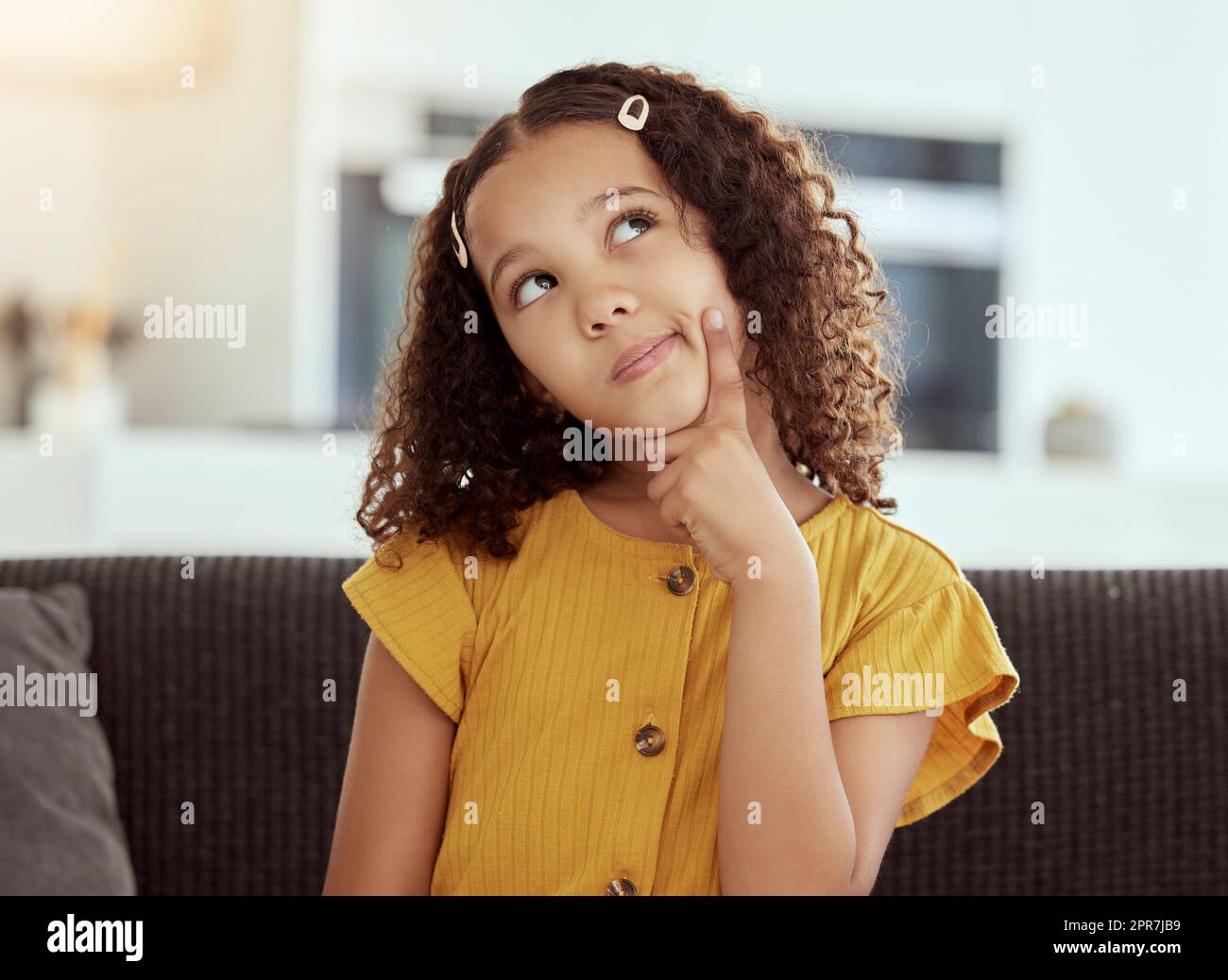 Adorable little mixed race child thinking at home. One small cute hispanic girl sitting alone on a sofa in a living room and coming up with an idea. Bored young kid with curly hair with a solution Stock Photo