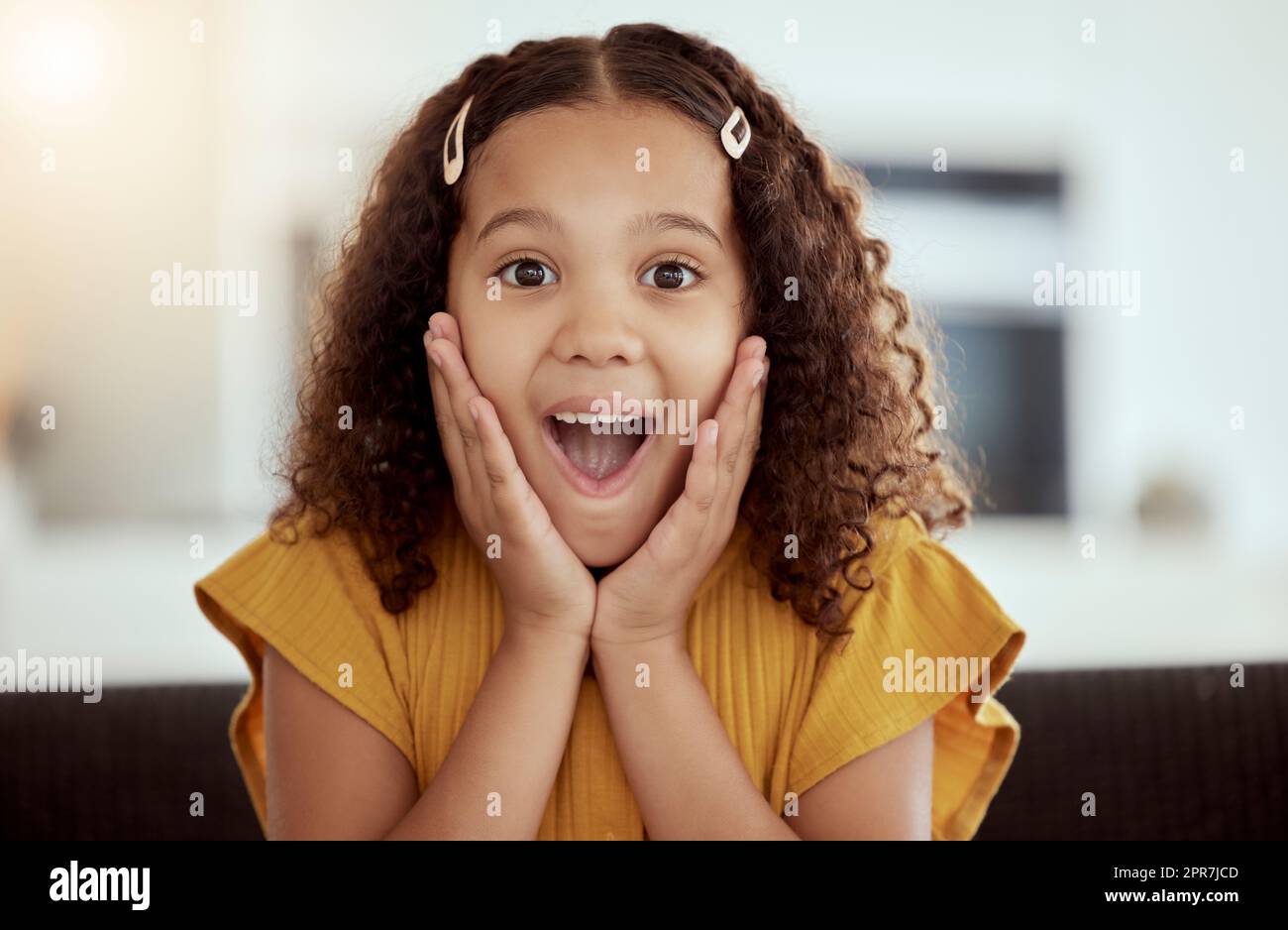 Adorable little mixed race child touching face with her hands in surprise at home. One small cute hispanic girl sitting alone on a living room sofa and looking shocked. Excited kid with curly hair Stock Photo