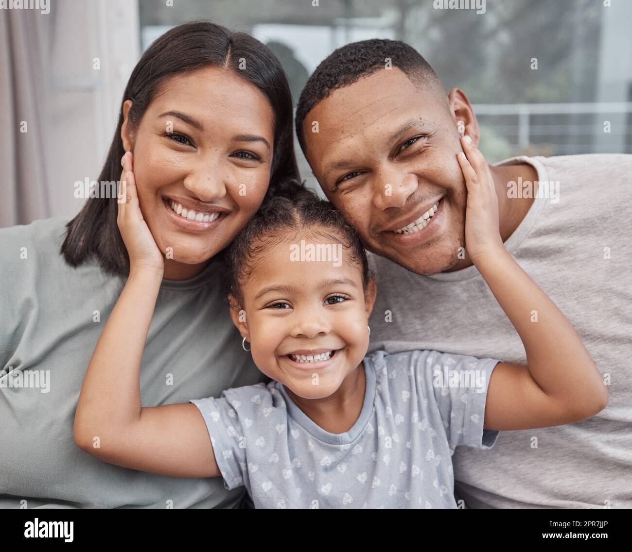 Happy and affectionate young mixed race family of three sitting on a sofa in the living room at home. Married couple sitting with their cute little girl in the lounge. Mom, dad and adorable daughter Stock Photo