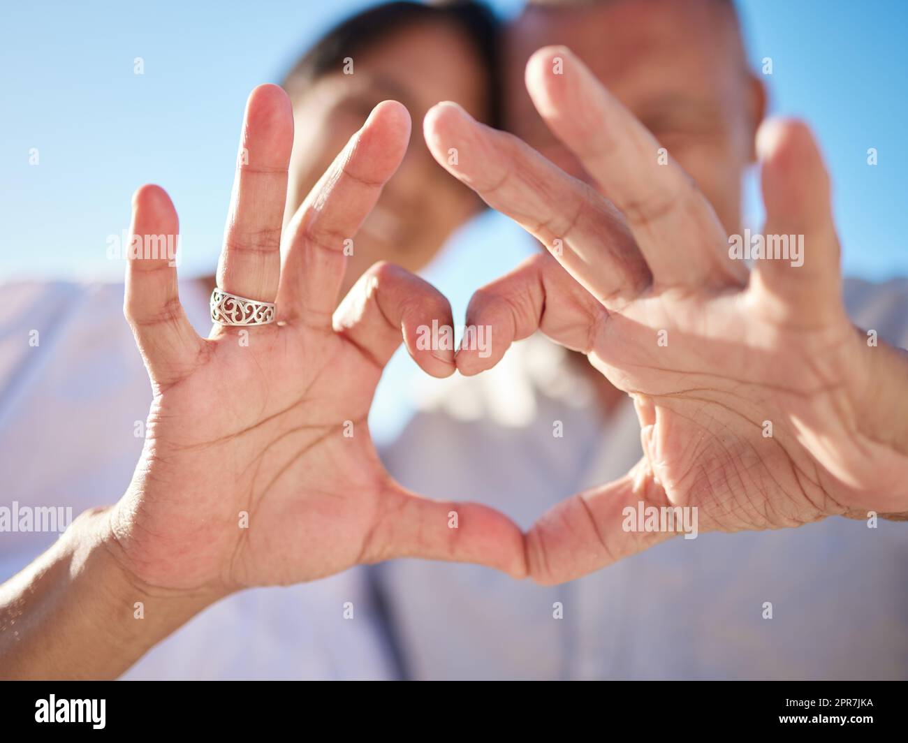 Affectionate mature mixed race couple sharing an intimate moment on the beach. Senior husband and wife making a heart shape with their hands. They love spending time together by the sea at sunset Stock Photo