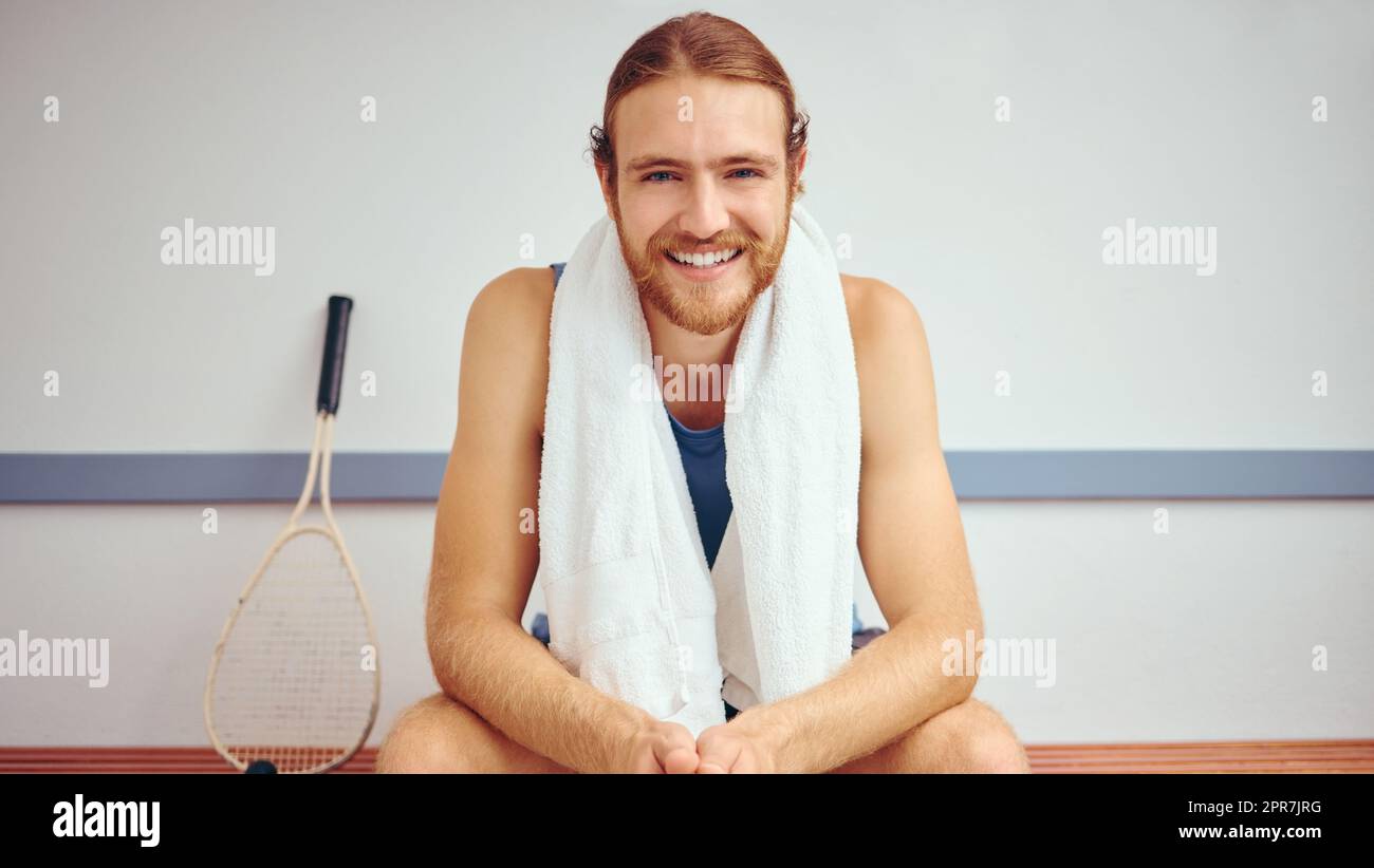 Portrait of a squash player with his racket. Caucasian man sitting in a gym locker room relaxing. Fit, young athlete taking a break before his match. Happy healthy player sitting in his gym Stock Photo