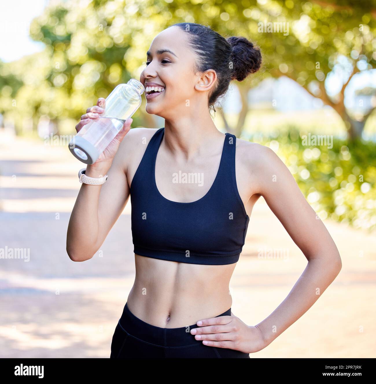 One fit young mixed race woman taking a rest break to drink water from bottle while exercising outdoors. Happy female athlete quenching thirst and cooling down after running and training workout Stock Photo