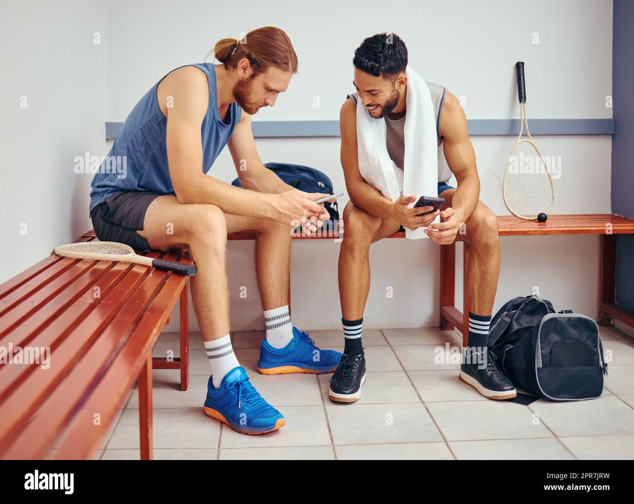 Two players bonding in their gym locker room. Two professional athletes using their cellphones together. Professional squash players talking and using their smartphones to text in the locker room Stock Photo