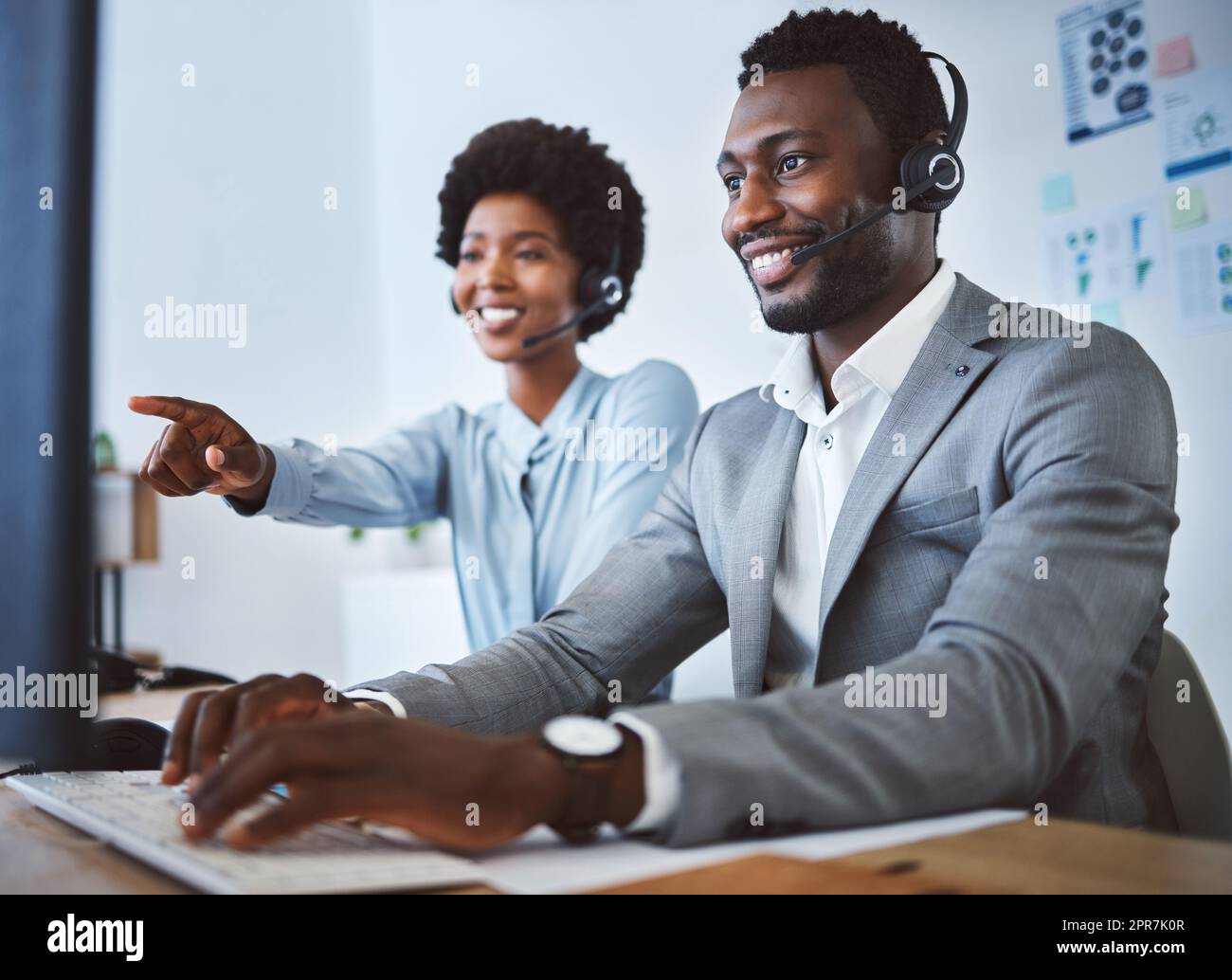 Happy african american male call centre telemarketing agent discussing plans with colleague while working together on computer in an office. Two consultants troubleshooting solution for customer service and sales support Stock Photo