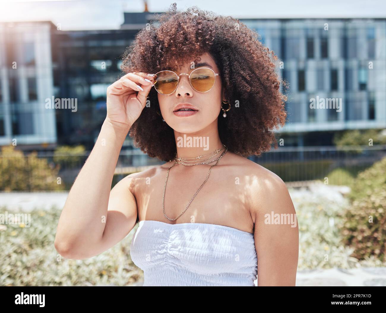 Portrait of young trendy beautiful mixed race woman with an afro smiling and posing alone outside. Hispanic woman wearing sunglasses and feeling happy. Fashionable african american woman in the city Stock Photo
