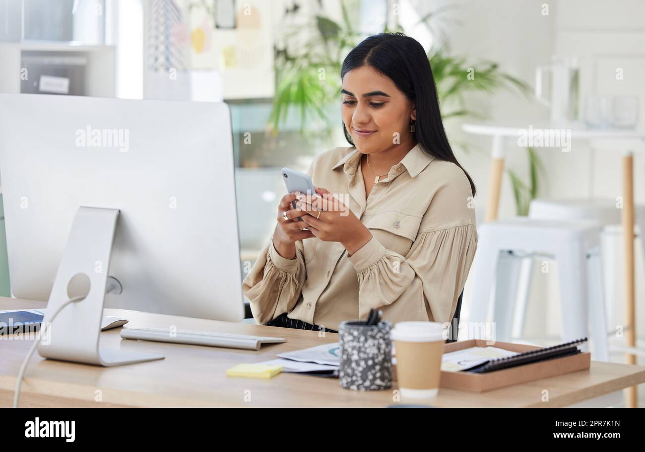 One beautiful young mixed race businesswoman using her smartphone during a lunch break in the office at work. Confident and successful female entrepreneur of indian ethnicity working on a cellphone in her workplace looking motivated and happy. Stock Photo