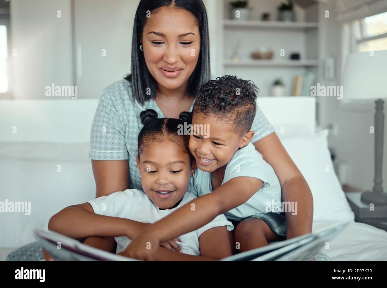 Young mixed race mother reading a storybook while relaxing at home with her two children. Smiling parent telling small kids funny fairy tale story kids pointing at book while they sit together Stock Photo