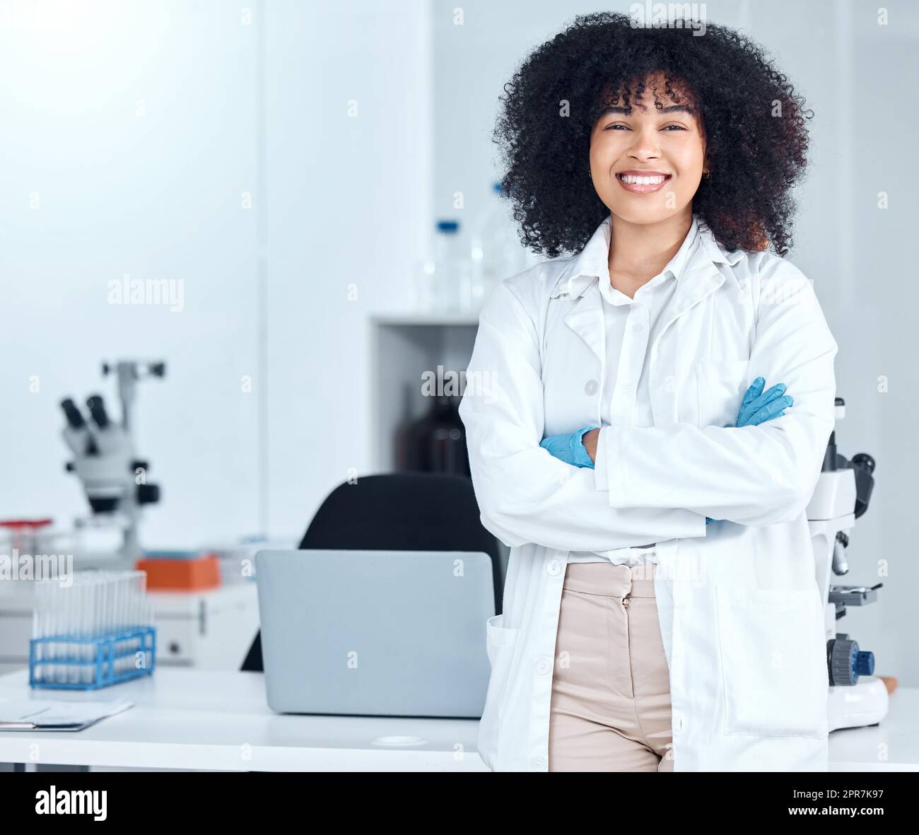 Portrait of a beautiful young african american woman with an afro wearing a labcoat and gloves while standing with her arms crossed in the laboratory. A mixed race female scientist smiling happily Stock Photo