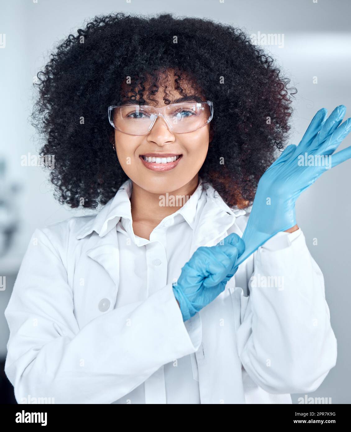 Portrait of young african american female scientist with afro hair wearing a labcoat and goggles while putting on gloves in the laboratory. A mixed race female scientist getting ready to conduct an experiment Stock Photo