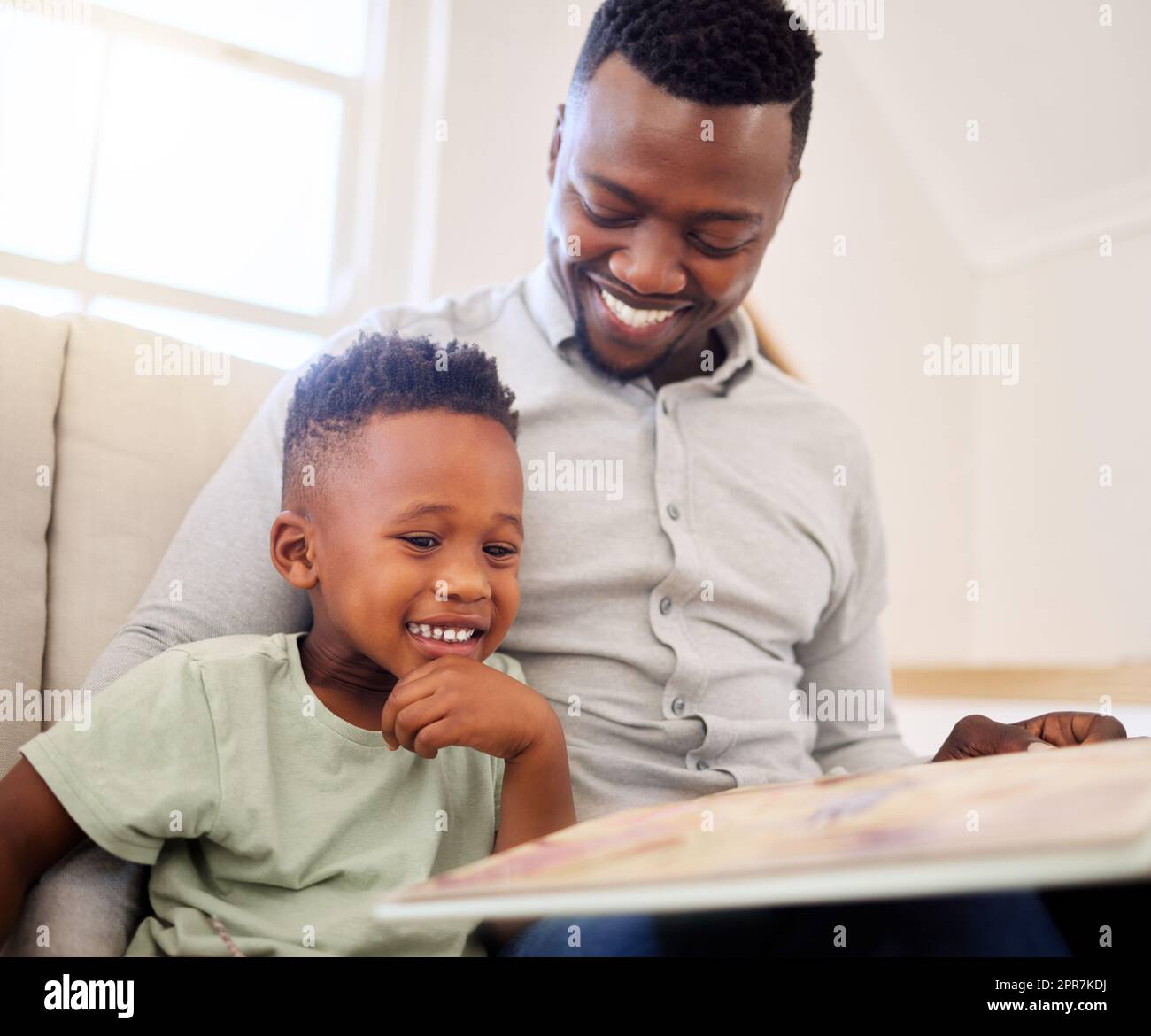 African american father bonding with his at home. Black male helping his son read a book and practice learning while sitting on a sofa at home. cute little black boy reading to his dad in the lounge. father and son smiling while learning some educational Stock Photo