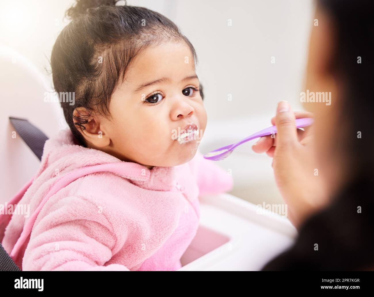 One mixed race baby girl being fed with a spoon by mom at home. Cute toddler with her mouth stuffed with porridge sitting in a feeding chair to enjoy some soft foods. Nutrition for healthy development Stock Photo