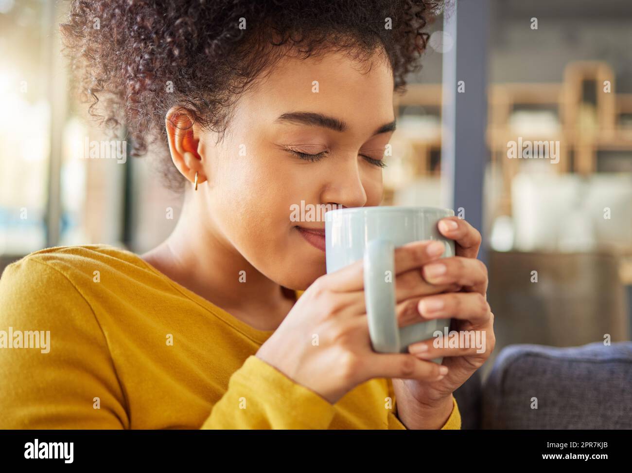 Young happy mixed race woman holding and drinking a cup of coffee at home. One cozy hispanic female smelling and enjoying a cup of tea while relaxing at home Stock Photo