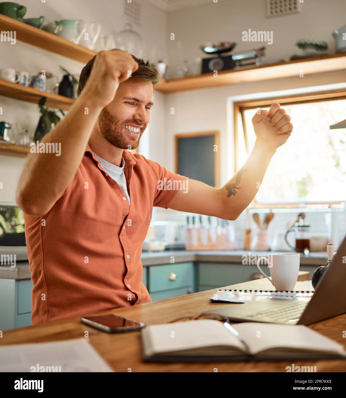 Young cheerful caucasian businessman cheering in success while working on a laptop at home alone. One joyful male businessperson smiling while working in the kitchen at home Stock Photo
