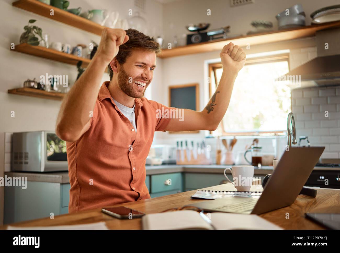 Young cheerful caucasian businessman cheering with joy while working on a laptop at home alone. One joyful male businessperson smiling while working in the kitchen at home Stock Photo