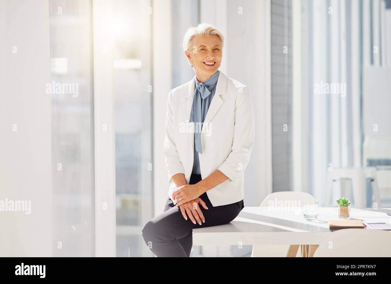 Portrait of a confident mature caucasian businesswoman sitting on a table in an office alone. One female only with grey hair smiling and looking cheerful. Ambitious entrepreneur and determined leader ready for success in her startup Stock Photo