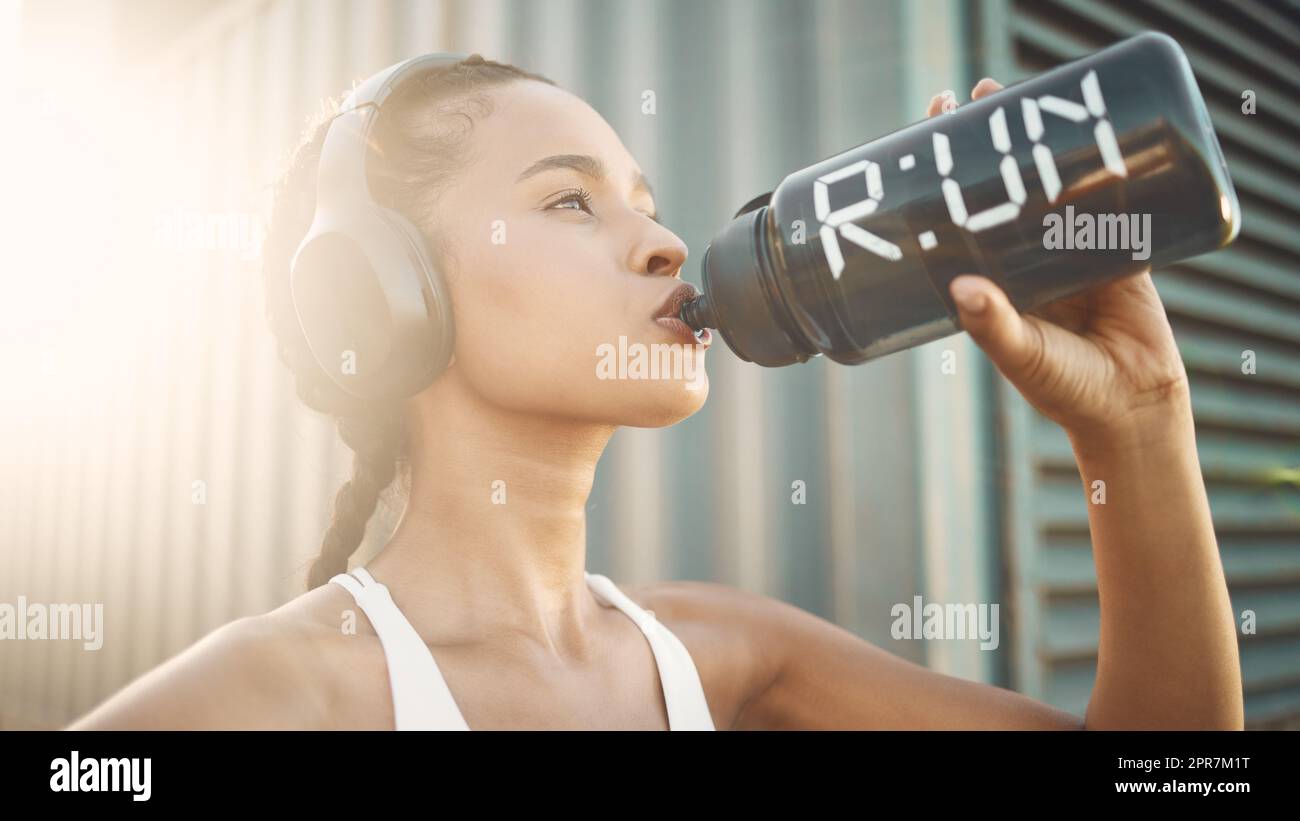 One fit young hispanic woman wearing headphones and taking a rest break to drink water from a bottle while exercising outdoors. Female athlete quenching thirst and cooling down after run and training workout in an urban setting Stock Photo