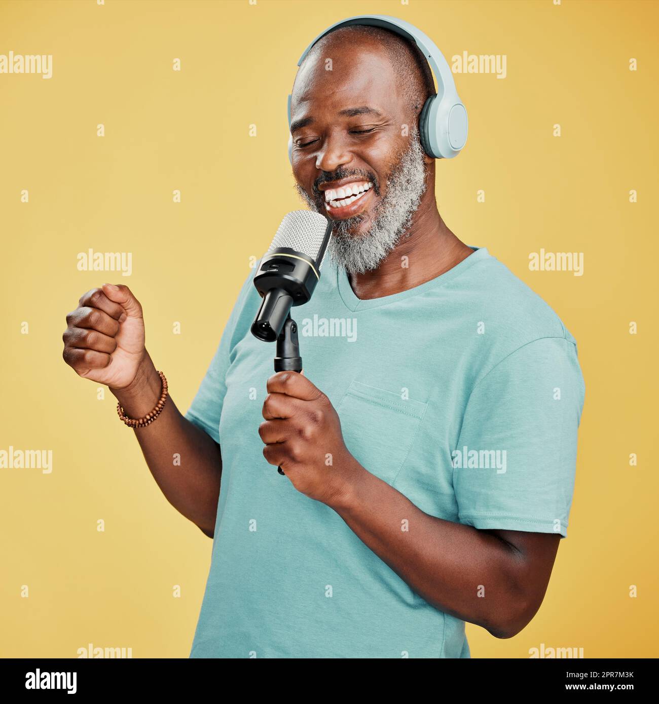 Happy mature African American man against a yellow studio background wearing headphones to listen to music while singing karaoke with a microphone. Smiling black man enjoying and holding a mic to sing Stock Photo