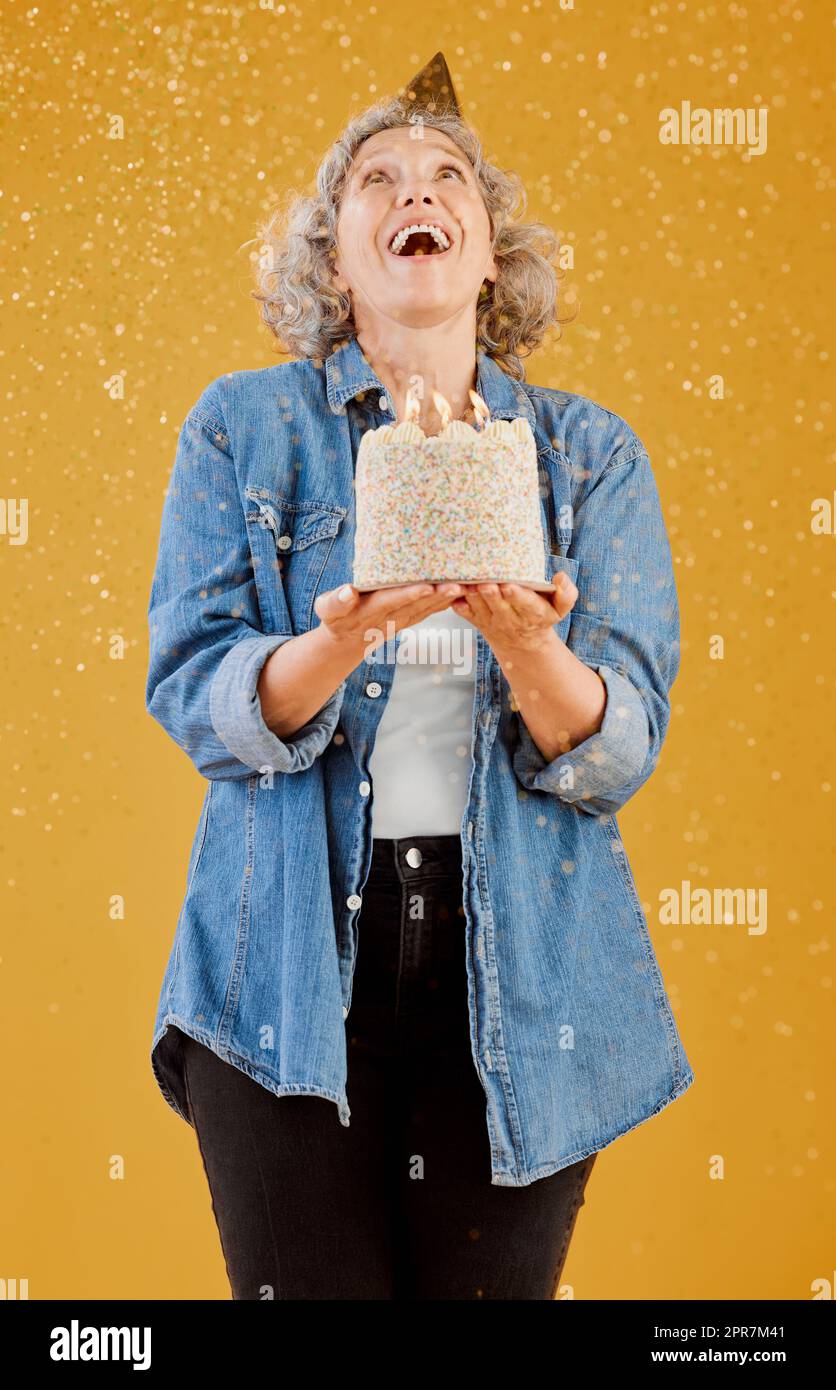One happy mature caucasian woman wearing a birthday hat and holding a cake while confetti falls from above against a yellow background in the studio. Smiling white lady celebrating another year Stock Photo