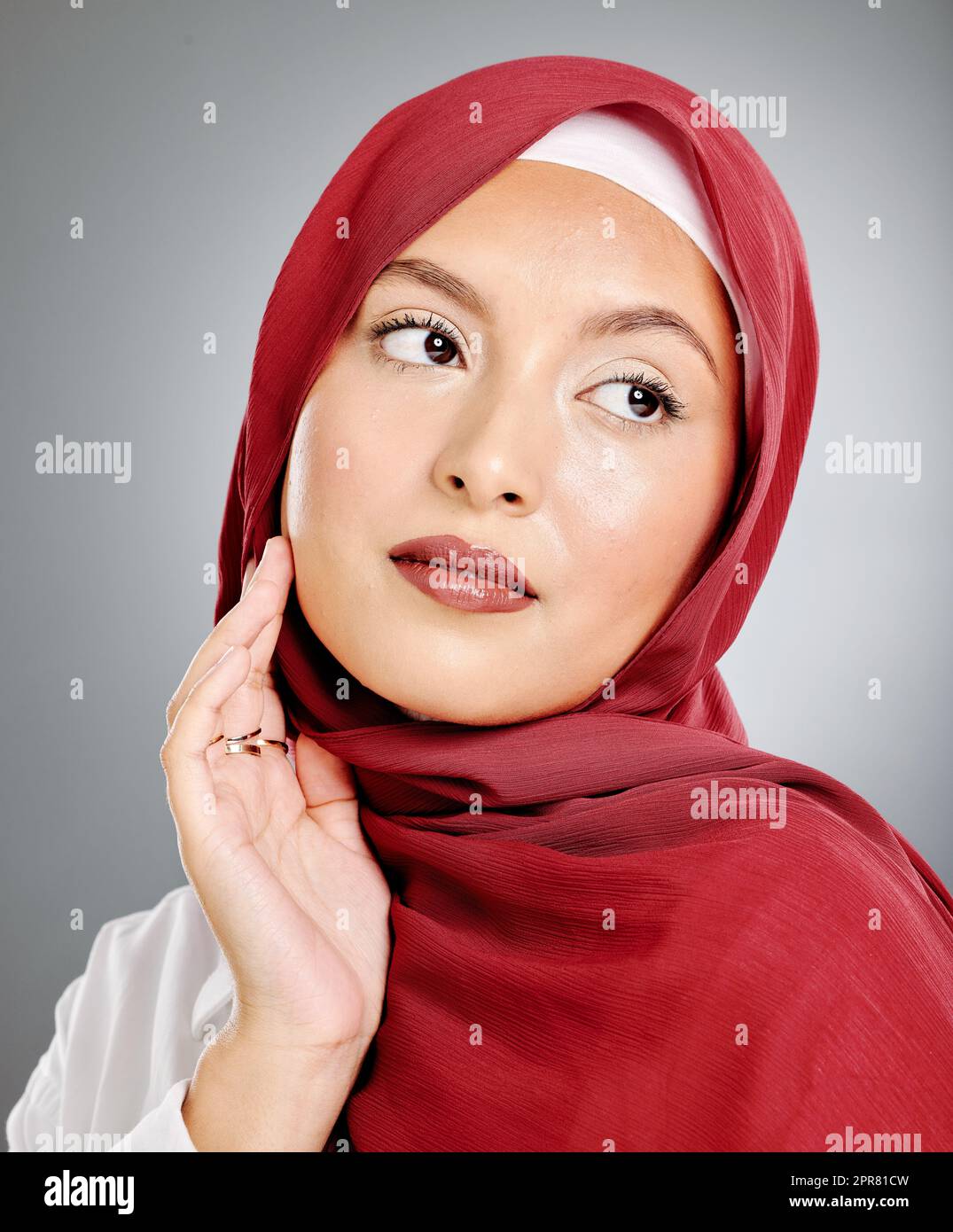 A glowing beautiful muslim woman isolated against grey copyspace background. Young woman wearing a hijab or headscarf showing her eyelash extensions while daydreaming, touching her flawless skin Stock Photo