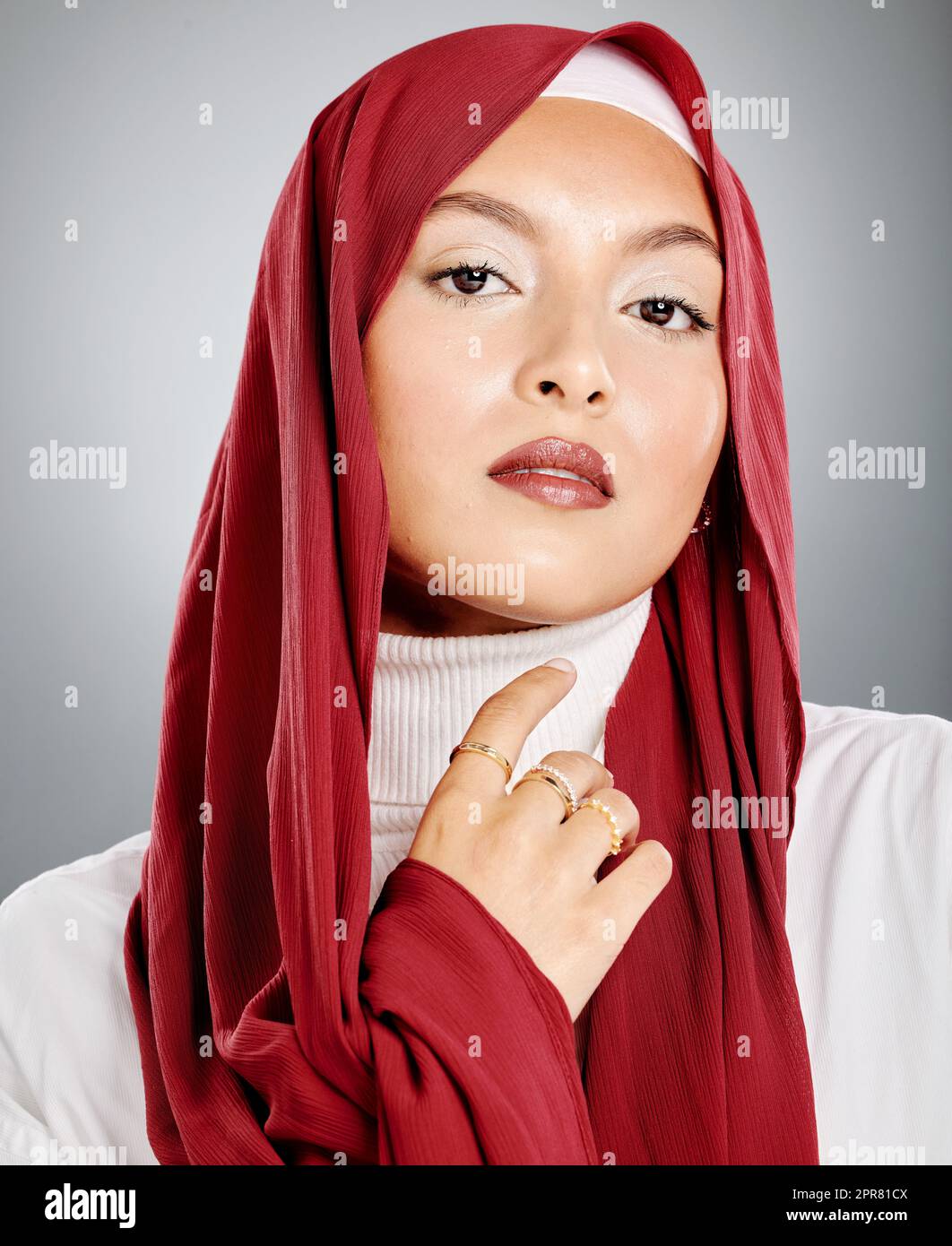 Portrait of a Muslim woman wearing a red hijab or headscarf showing her eyelash extensions and makeup. Showing her flawless skin glowing. Beautiful woman isolated against a grey studio background. Stock Photo