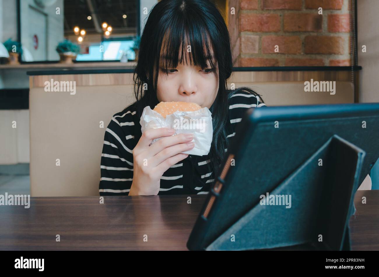 young girl is eating a hamburger and watching an online social media tech tablet on the table. Stock Photo